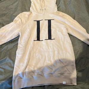 Beige hoodie från Les Deux med tryck - Säljer en beige hoodie från Les Deux med stor svart II-tryck på bröstet. Tröjan har huva utan snörning, känguruficka och ribbade muddar. Perfekt för dig som gillar stilren och avslappnad look.