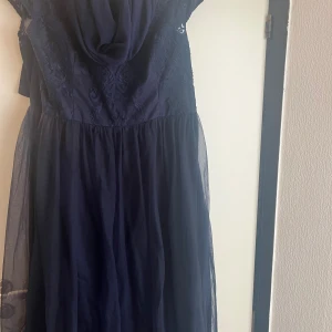 Marinblå långklänning med spets - Elegant marinblå långklänning med vackert spetsliv och transparent mesh i kjolen. Klänningen har korta ärmar och en rundad halsringning med fina spetsdetaljer upptill. Kjolen är lätt och faller snyggt, perfekt för speciella tillfällen. Det tillkommer en sjal med 