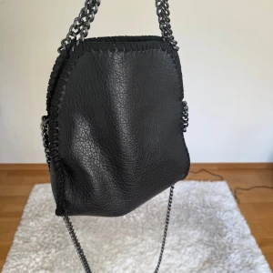 Svart axelväska med kedjerem - Cool svart axelväska i skinnimitation med grov kedjerem i metall och dekorativa sömmar längs kanterna. Väskan har en mjuk, något slouchy form och stängs med magnetknapp. Perfekt för dig som gillar en edgy och stilren look.