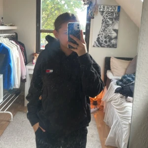 Svart hoodie Tommy Jeans XL - Svart hoodie från Tommy Jeans i storlek XL. Klassisk modell med huva och snörning, samt broderad logga på bröstet i rött, vitt och blått. 