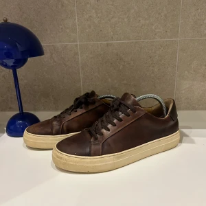 Bruna sneakers från Sneaky Steve - Snygga bruna sneakers från Sneaky Steve i skinn med klassisk vit sula. Skorna har rund tå, platt sula och snörning. Perfekta för dig som gillar stilrena och tidlösa sneakers med en touch av vintagekänsla.
