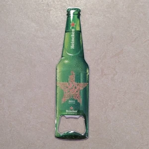 Heineken Music Edition flasköppnare / magnet - Snygg flasköppnare och magnet i form av en Heineken-flaska, Music Edition. Tillverkad i metall med tryckt design. Perfekt för baren eller som samlarobjekt eller som kylskåpsmagnet. Gott skick, endast mindre bruksspår synliga.