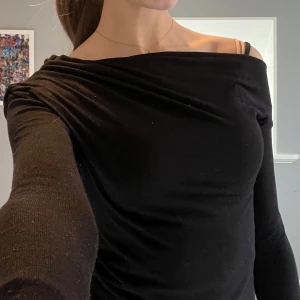 Svart offshoulder topp från Nelly - Säljer en svart långärmad topp från Nelly med offshoulder-design. Toppen har en snygg drapering över axlarna och är figurnära. 