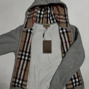 Grå hoodie från Burberry  - Snygg grå hoodie från Burberry med klassiskt rutigt foder i beige, svart och rött. Tröjan har huva med snörning, silverfärgad dragkedja och broderad Burberry-logga på bröstet. Perfekt för dig som gillar stilrena detaljer och exklusiva märken. Skickas imon 24 timmar!