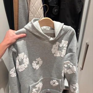 Denim Tears Hoodie - Säljer en grå hoodie från Denim Tears med vita blommiga applikationer över hela tröjan. Tröjan har huva, magficka och långa ärmar. Perfekt för dig som vill sticka ut med en unik och trendig look.