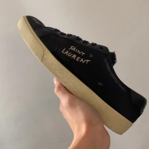 Saint Laurent skor - Svarta skor från Saint Laurent med handskriven logga på sidan. Skorna har slitna detaljer, svarta snören och beige sula. Ovandelen är i canvas och läder, med råa kanter för en edgy look. Perfekta för dig som vill ha skor som passar för alla tillfällen. Skick 8/10. Man får även med ett par extra skosnören i en Saint Laurent dust bag.