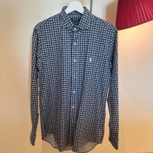 Mönstrad skjorta från Polo Ralph Lauren - Snygg skjorta från Polo Ralph Lauren i custom fit. Skjortan är mörkblå med vitt rutmönster och har klassisk krage samt knappar framtill. Tillverkad i mjuk bomull och har långa ärmar. Perfekt för dig som gillar stilrena detaljer och mönster.