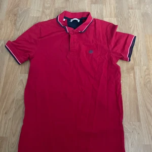 Röd pikétröja Selected Homme M - Snygg röd pikétröja från Selected Homme med broderad logga på bröstet. Tröjan har klassisk krage med mörkblå och vita ränder samt korta ärmar med matchande kant. Tillverkad i ekologisk bomull för skön känsla och stilren look.