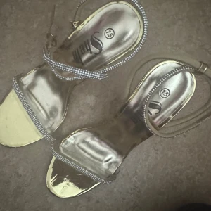 Guldiga sandaler med strass och klack - Säljer ett par glammiga sandaler från Shein i metallic guld med smala remmar dekorerade med strass. Skorna har öppen tå, hög klack och transparent detalj framtill. Perfekta för dig som vill sticka ut och glänsa.