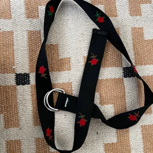 Svart tygbälte med broderade rosor - Svart tygbälte med broderade röda rosor och gröna blad längs hela bandet. Bältet har en silverfärgad dubbel D-ring som spänne och är gjort i ett mjukt, flexibelt material. Perfekt accessoar för att ge outfiten en cool och personlig touch.