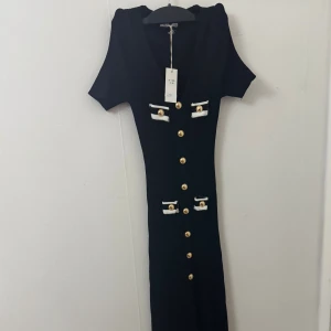 Svart långklänning från Giorgia - Supersnygg elegant svart långklänning från Giorgia med korta ärmar och djup v-ringning. Klänningen har dekorativa guldknappar framtill och vita detaljer vid fickorna. Ribbad struktur som ger en tight och smickrande passform. Perfekt för dig som vill sticka ut med stilrena detaljer. Storlek S/M, den är strechig och i bra och tjockt material. Nyspris 699 kr 