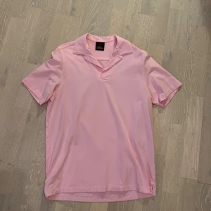 Ljusrosa piké Oscar Jacobson - Säljer en stilren ljusrosa skjorta från Oscar Jacobson i mjuk bomull. Skjortan har korta ärmar, öppen krage och relaxed fit. Perfekt för dig som vill ha en clean och fräsch look med en touch av färg.