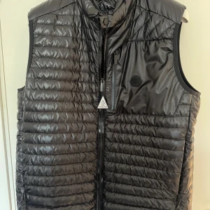 Moncler Aptera Down Vest Black Edition - Snygg svart dunväst från Moncler med quiltad design och hög krage. Västen har dragkedja framtill, en bröstficka med dragkedja och diskret Moncler-logga på bröstet. Perfekt för lager-på-lager och streetstyle.
