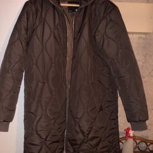 Quiltad brun jacka "Diana" - Säljer en lång, quiltad brun jacka "Diana" med dragkedja framtill och ribbade muddar vid ärmsluten. Jackan har en oversized passform och är perfekt för kyligare dagar. Snyggt mönstrad med vågformade sömmar och tillverkad i syntetmaterial.
