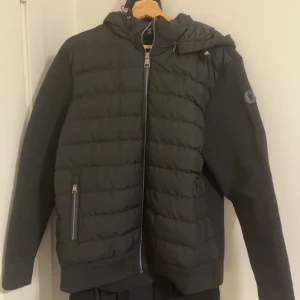 Svart pufferjacka från Moncler - Snygg svart pufferjacka från Moncler storlek XL med stickade ärmar och huva. Jackan har dragkedja framtill, två fickor med dragkedja och Moncler-logga på ärmen. Perfekt för kyliga dagar och ger en clean, modern look.