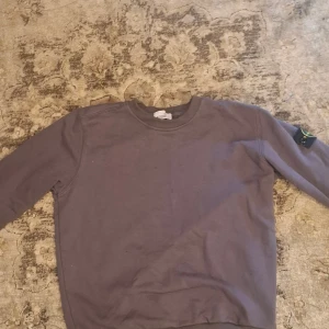 Grå sweatshirt från Stone Island - Säljer en grå sweatshirt från Stone Island med klassisk rund hals och ribbade muddar. Tröjan har den ikoniska Stone Island-loggan på vänster ärm och är tillverkad i mjuk bomull. Perfekt för dig som gillar streetwear och stilren design.