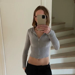 Grå croppad kofta med dragkedja - Säljer en grå croppad kofta med dragkedja framtill och huva. Tröjan har ribbade detaljer på ärmar och midja, samt lång ärm. Perfekt för dig som gillar en trendig och avslappnad stil.💕☺️