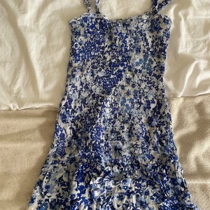 Blåvit blommig kortklänning från ONLY - Supersöt kortklänning från ONLY med blått och vitt blommönster, smockad överdel och volangdetaljer nertill. Klänningen har smala axelband och är perfekt för varma dagar. Materialet känns lätt och luftigt.