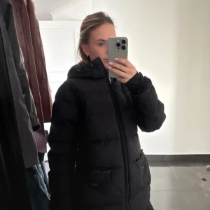 Svart pufferjacka med huva - Svart pufferjacka med huva och dragkedja framtill. Jackan har två stora fickor med lock och en enkel, stilren design. Perfekt för kalla dagar och ger en skön, oversized känsla. Passar dig som gillar en klassisk look med streetvibe.