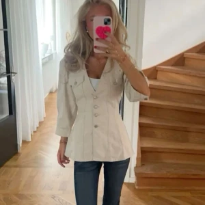 Beige jeansjacka från Avora med knappar - Snygg beige overshirt från Avora med markerad midja, v-ringning och knappar framtill. Jackan har trekvartsärmar, två bröstfickor med lock och justerbara detaljer i ryggen. 