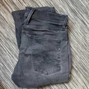 Replay Anbass svarta jeans, stl 31 - Svarta Replay Anbass jeans i slim fit med klassisk femficksdesign. Jeansen har en mörk tvätt, raka ben och är tillverkade i stretchig bomullsblandning för extra komfort. Perfekta för dig som gillar en stilren och modern look. En defekt mellan benan men syns inte i användning pga färgen