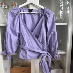 Ljuslila omlottblus med knyt - Supersöt ljuslila blus från LINDEX med omlottmodell och knyt i midjan. Blusen har puffiga långärmar med resår vid ärmslut och en djup v-ringning. Perfekt för dig som gillar trendiga och feminina detaljer.