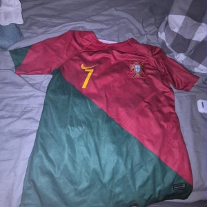 Portugal Ronaldo #7 Nike fotbollströja - Säljer en Portugal fotbollströja från Nike med Ronaldo och nummer 7 på ryggen. Tröjan har korta ärmar, diagonal design i vinrött och mörkgrönt, samt gula detaljer. Materialet är Dri-FIT polyester, perfekt för träning eller match. Landslagsemblem på bröstet.