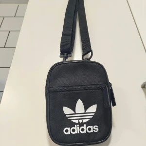 Svart axelväska från Adidas - Snygg svart axelväska från Adidas med loggan framtill. Väskan är kompakt, har justerbar axelrem och dragkedja både på huvudfacket och framfickan. Perfekt för mobilen, plånboken och andra småsaker. Tillverkad i slitstark polyester.