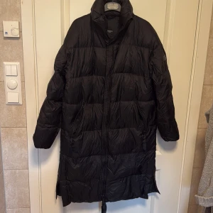 Svart lång dunjacka Rockandblue - Lång svart dunjacka från Rockandblue med hög krage och dragkedja framtill. Jackan har två fickor med dragkedja, slitsar med knappar i sidorna och quiltad design. Perfekt för kalla dagar och riktigt snygg till streetwear.