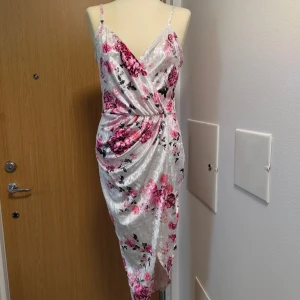 Blommig omlottklänning från NLY One - Snygg omlottklänning från NLY One i glansigt material med blommigt mönster i rosa och svart på vit botten. Klänningen har smala axelband, v-ringning och draperad midja. Perfekt för dig som vill sticka ut med en feminin och trendig look.