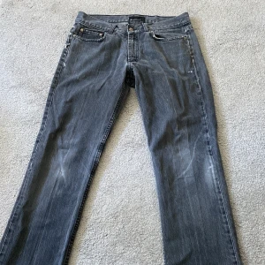 Grå jeans från Acne Action Jeans - Säljer ett par grå jeans från Acne Action Jeans med klassisk femficksdesign och raka ben. Jeansen har en mörkgrå tvätt med subtila slitningar och är tillverkade i denim. Perfekta för dig som gillar en stilren och tidlös look.