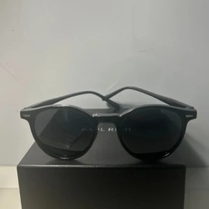 Svarta runda solglasögon  - Snygga svarta solglasögon med rundad form och klassisk design. Bågarna är i svart plast med diskreta silvriga detaljer vid gångjärnen. Perfekta för att lyfta din stil under soliga dagar.