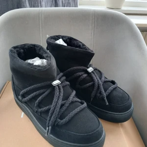 Nya Inuikii boots strl 39 - Säljer ett par svarta Inuikii boots med grova snören och mjuk fuskpäls på insidan. Skorna är i mocka och har en platt sula med Inuikii-logga baktill och undertill. Perfekta för kalla dagar och riktigt snygga till vinterlooken.