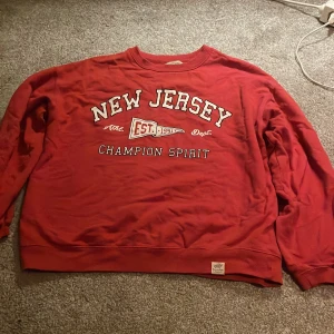 Röd sweatshirt New Jersey - Röd sweatshirt med tryck 'New Jersey Champion Spirit' och detaljer i vitt. Rund halsringning och långa ärmar. Perfekt för dig som gillar college-stil och vill ha en skön tröja till vardags.