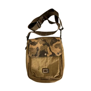 Guess crossbody väska  - Cool crossbody väska från Guess i beige, grön och brunt med camouflage-mönstrat lock. Väskan har justerbar axelrem, flera fack och detaljer i skinn och metall. Perfekt för dig som vill ha en streetig och praktisk väska med snygga detaljer. Massa små fickor överallt så det finns plats för mer än man tror😻🙏🏻