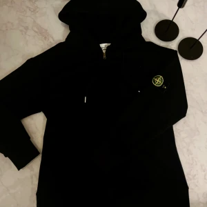 Svart hoodie från Stone Island M - Svart hoodie från Stone Island med klassisk logga på ärmen. Tröjan har huva med snörning och dragkedja framtill. Perfekt för dig som gillar streetwear och vill ha en stilren look.