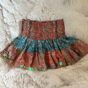 Färgglad blommig kortkjol från Ananda Wild - Supersöt kortkjol från Ananda Wild med blommigt mönster i rött, grönt, blått och brunt. Kjolen har smockad midja och volangig nederdel, perfekt för sommarens alla äventyr. Lätt och luftig i bomull, med fina detaljer och färgskiftningar.