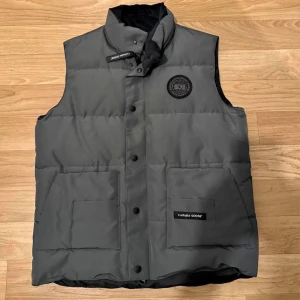 Grå dunväst från Canada Goose - Snygg grå dunväst från Canada Goose med hög krage och svarta tryckknappar. Västen har två stora fickor framtill, diskret logga på bröstet och innerfoder i svart satin. Perfekt för kalla dagar när du vill hålla stilen.