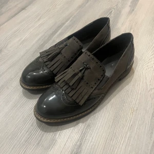 Bruna loafers med fransar och tofsar - Snygga bruna loafers med dekorativa fransar och tofsar framtill. Skorna har en mix av blankt och matt material, broguedetaljer och en platt sula med markerad söm runt om. Perfekta för dig som vill ha en klassisk men trendig look.