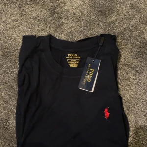Svart t-shirt från Polo Ralph Lauren - Svart t-shirt från Polo Ralph Lauren med röd broderad logga på bröstet. Klassisk rund halsringning och korta ärmar. Slim fit-modell i mjuk bomull, perfekt för en clean och stilren look.
