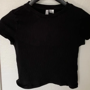 Svart ribbad croppad topp H&M Divided - En svart, ribbad croppad topp från H&M Divided. T-shirten har rund halsringning och korta ärmar. Enkel och stilren modell som är lätt att matcha med allt. Perfekt för dig som gillar en basic look med en twist.