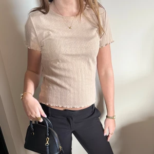 Beige ribbad kortärmad topp - Säljer en beige ribbad topp med korta ärmar och diskret mönster. Toppen har en lätt vågig kant nertill och vid ärmslut, samt rund halsringning. Perfekt för dig som gillar en enkel men ändå söt stil.