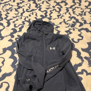 Blå vindjacka från Under Armour - Säljer en blå vindjacka från Under Armour med huva och hel dragkedja. Jackan har vita loggor på bröstet och ärmen, samt ventilerande meshpaneler på sidorna. Perfekt för utomhusaktiviteter och träning. Snygg och sportig look!