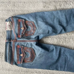True Religion Joey Low Rise Flare Jeans - Säljer ett par ljusblå True Religion Joey jeans med låg midja och bootcut/flare passform. Jeansen har tydliga orange kontrastsömmar, dekorativa ficklock med knappar och klassisk True Religion-logga bak. Tillverkade i jeansmaterial med snygg tvätt och detaljer.