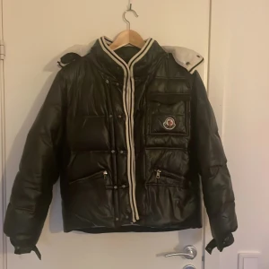 Svart pufferjacka från Moncler - Säljer en svart pufferjacka från Moncler med vita och röda detaljer. Jackan har dragkedja, knappar, flera fickor och en vit huva med ränder. Klassisk Moncler-logga på bröstet och coolt innerfoder med serietidningsmönster.