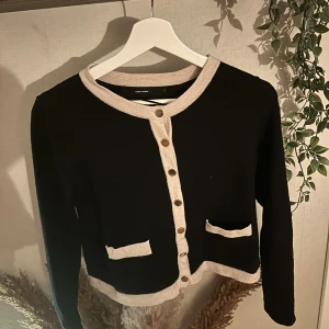 Svart kofta med beige detaljer Vero Moda - Stilren svart kofta från Vero Moda med beige kantband och dekorativa knappar framtill. Koftan har två små fickor fram och är långärmad. Perfekt för lager-på-lager och ger en chic vibe till din outfit.