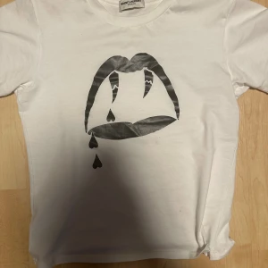 Hedi Slimane X Ysl Bloodluster - Snygg vit t-shirt från Saint Laurent med ett svart grafiskt tryck av läppar och tårar på framsidan. Klassisk rund hals och korta ärmar. Tillverkad i mjuk bomull som känns skön mot huden. Perfekt för dig som gillar statement-plagg och designer.