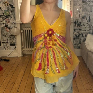 Gul linnetopp med blommor och paljetter - Unik gul linnetopp med v-ringning och blommiga detaljer framtill. Toppen har paljetter i rosa och grönt, broderade blommor och ett brett band i midjan som knyts bak. Materialet är lätt och luftigt, perfekt för att sticka ut med färg och mönster. jag vet inte vart den är köpt då jag fick den av en släkting men den passar xs-s