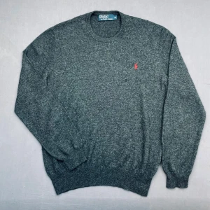 Grå stickad merinoull tröja från Polo Ralph Lauren - Mörkgrå stickad tröja från Polo Ralph Lauren med klassisk röd broderad logga på bröstet. Tröjan har rund halsringning och långa ärmar med ribbade muddar och nederkant. Det har funnits hål på tröjan men har blivit lagat hos en skräddare  