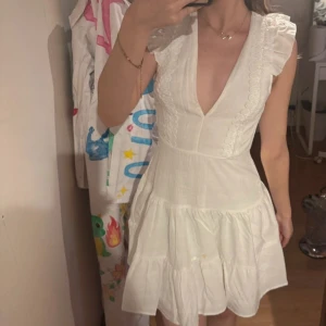 Vit kort klänning med volang och knyt - Studentklänning med djup v-ringning, volangdetaljer vid axlarna och knyt i ryggen. Super bra skick, har aldrig använt den. 